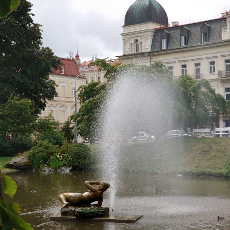 Brunnen mit bronzener Skulptur und Wasserfontäne in einem Park hinter historischen Gebäuden.