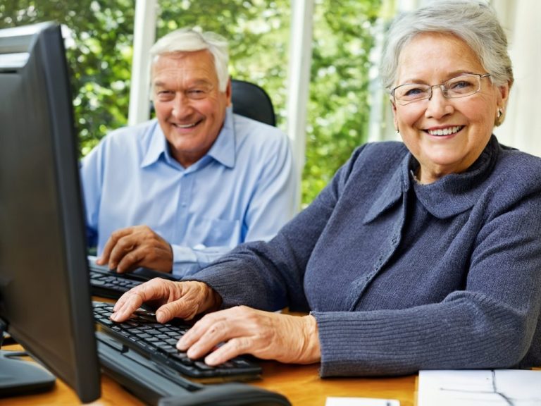 Ältere Frau und Mann lächeln, während sie an Computern arbeiten.