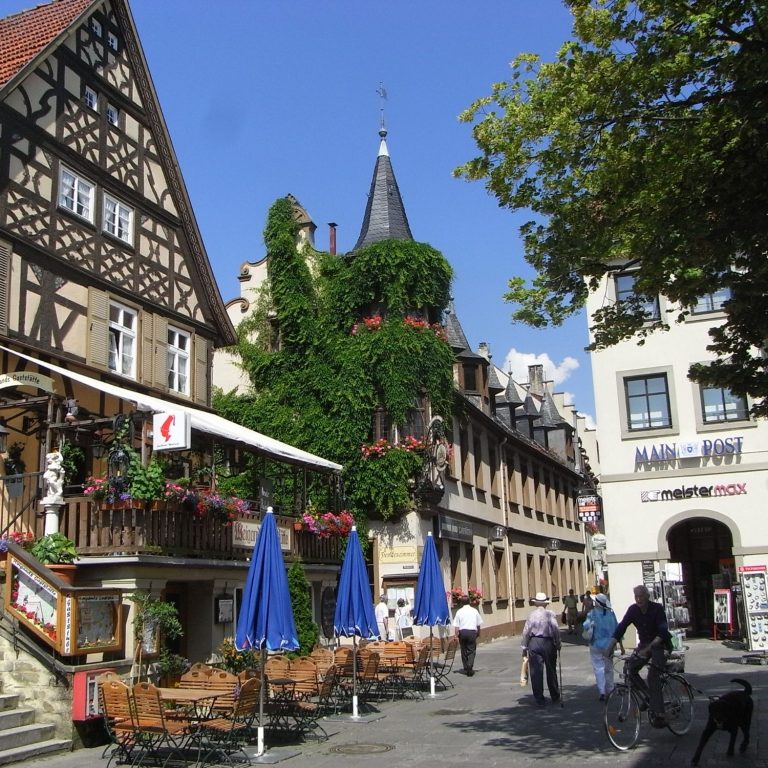 Gemütlicher Stadtplatz mit historischen Gebäuden, Blumenbalkonen und sonnigem Himmel.