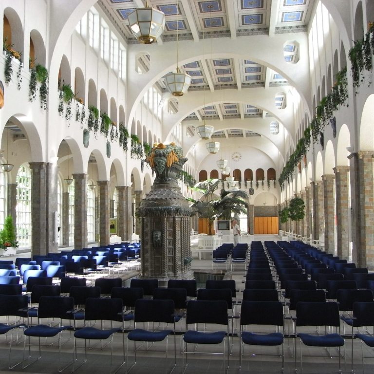 Großer Saal mit hohen Bögen, Reihen von Stühlen und Pflanzen an den Fenstern.